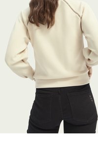 Felpa crema con design della parte posteriore testurizzato, vestibilità morbida e polsini a costine, abbinata a jeans neri con tasche posteriori.