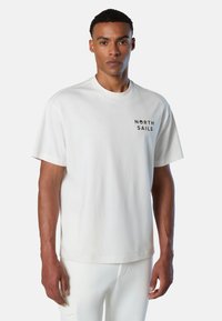Hombre vistiendo una camiseta blanca sencilla con el texto "NORTH SAILS" en el pecho y pantalones blancos a juego, de pie frente a un fondo blanco.