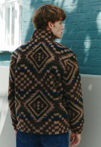 Brown fleece jacket with geometric diamond and check patterns in dark blue and green. High collar and relaxed fit; textured surface.