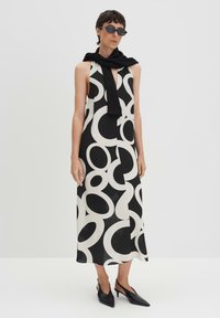 Schwarz-weißes ärmelloses Kleid mit kreisförmigen Mustern, aus leichtem Stoff. Kombiniert mit einem schwarzen Pullover, der um den Hals gebunden ist, und schwarzen High Heels.