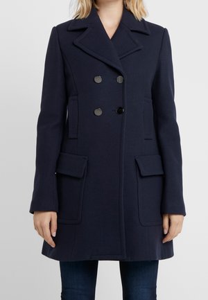 Femme portant un manteau bleu marine cintré à double boutonnage avec un grand col et des poches à rabat, associé à un jean bleu foncé.
