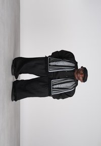 Άνδρας φορεμένος σε μαύρη στολή με φαρδιά παντελόνια, μακρύ μαντήλι που γράφει "HAN KJØBENHAVN," μαύρο καπέλο, στέκεται μπροστά σε απλό φωτεινό φόντο.