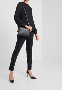 Schwarze plissierte Bluse mit hohem Kragen, kombiniert mit maßgeschneiderten schwarzen Hosen und einer strukturierten schwarzen Umhängetasche mit Kettenriemen.