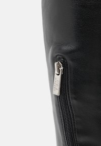 Botín de cuero negro con cremallera lateral y una etiqueta metálica estampada con el nombre de la marca. Superficie texturizada y diseño sin costuras.