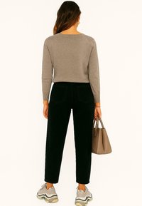 Grijze gebreide trui met lange mouwen, gecombineerd met hoge zwarte broeken. Model houdt een beige handtas vast en draagt grijze sneakers.