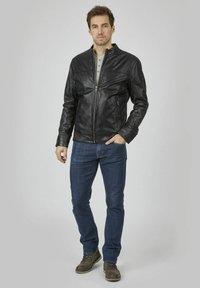 Schwarze Lederjacke mit einem Reißverschluss vorne, Stehkragen und taillierten Ärmeln, kombiniert mit blauen Jeans und braunen Stiefeln. Neutraler Hintergrund.