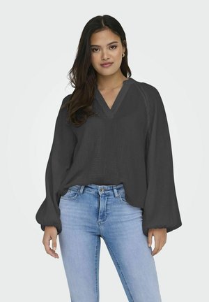 Blusa leggera di un grigio scuro, con scollo a V e maniche ampie e voluminose, abbinata a un paio di jeans aderenti blu chiaro. Tessuto liscio, design casual.