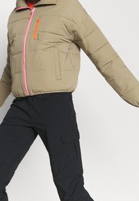 Giacca puffer beige con cerniera rosa, dettagli arancioni sulle tasche e colletto a coste. Abbinata a pantaloni cargo neri con tasche laterali.