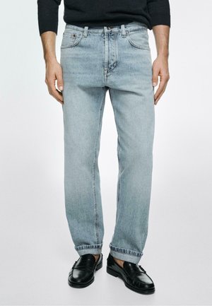 Mann trägt hellblaue Straight-Fit-Jeans mit aufgekrempelten Säumen und schwarzen Lederloafern, steht vor einem schlichten Hintergrund.