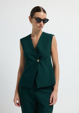 Green sleeveless blazer with a deep V-neck, gold button detail at the front, tailored shape, and smooth texture. Paired with matching pants.