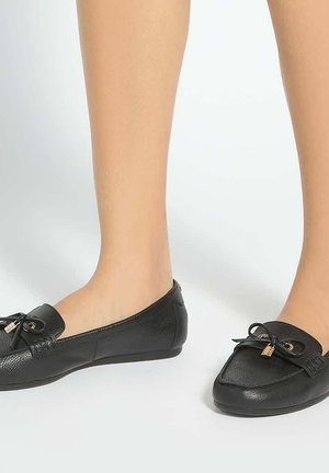 Beine, die schwarze Leder-Slipper mit schwarzen Schleifen und goldenen Akzenten auf hellem Hintergrund tragen.