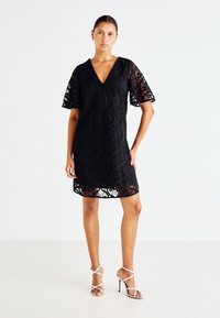 Μαύρο φόρεμα από δαντέλα με V-ντεκολτέ και κοντά φουσκωτά μανίκια, με περίπλοκο σχέδιο. Συνδυάζεται με ασημένιες ψηλές γόβες με λουριά.