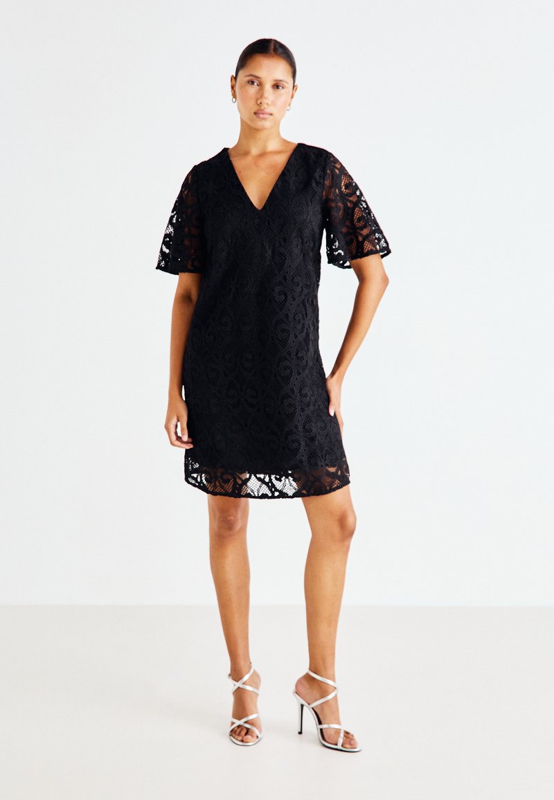 Μαύρο φόρεμα από δαντέλα με V-ντεκολτέ και κοντά φουσκωτά μανίκια, με περίπλοκο σχέδιο. Συνδυάζεται με ασημένιες ψηλές γόβες με λουριά.