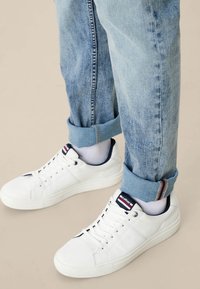 Zapatillas blancas con acabado de cuero texturizado, diseño con cordones y detalles en azul marino y rojo en la lengüeta. Combinadas con jeans de mezclilla claros y doblados.
