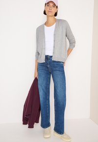 Cardigan grigio sopra una maglietta bianca, jeans blue straight-leg e sneakers beige. Tiene in mano una giacca bordeaux. Tessuti morbidi e stile casual.