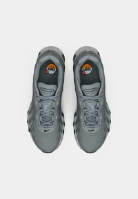 Light grey athletic shoes with a textured upper, rounded toe, and black accents on the sole. Features grey laces and visible branding.