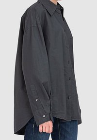 Personne portant une chemise ample à manches longues, boutonnée, gris foncé, col pointu et poche avant, associée à un jean bleu.