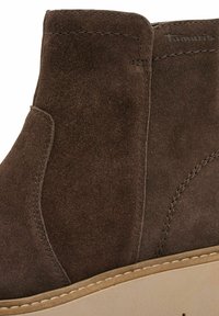 Bottine en daim marron avec un bout arrondi, des panneaux latéraux élastiques et une semelle en caoutchouc beige avec des accents de couture visibles.