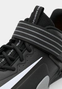 Chaussure de musculation noire avec un dessus texturé, un bout renforcé, un logo Nike blanc bien visible et une épaisse sangle noire avec des lignes réfléchissantes.