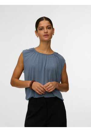 Ženska s temnimi lasmi v modri brezrokavnik srajci in črnih hlačah, z lesenim zapestnico, stoji pred svetlim enobarvnim ozadjem.