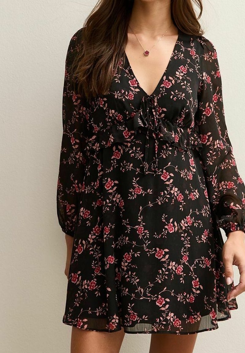 Femme portant une robe noire à fleurs avec des manches longues et un col en V, ornée de motifs de fleurs rouges et roses et une ceinture à la taille.