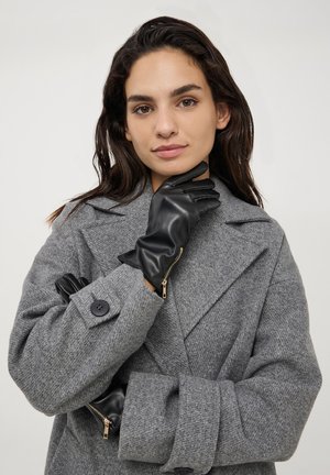 Guanti in pelle nera dal design elegante con dettagli in zip, indossati con un cappotto grigio testurizzato con ampie rever e chiusura a bottoni.