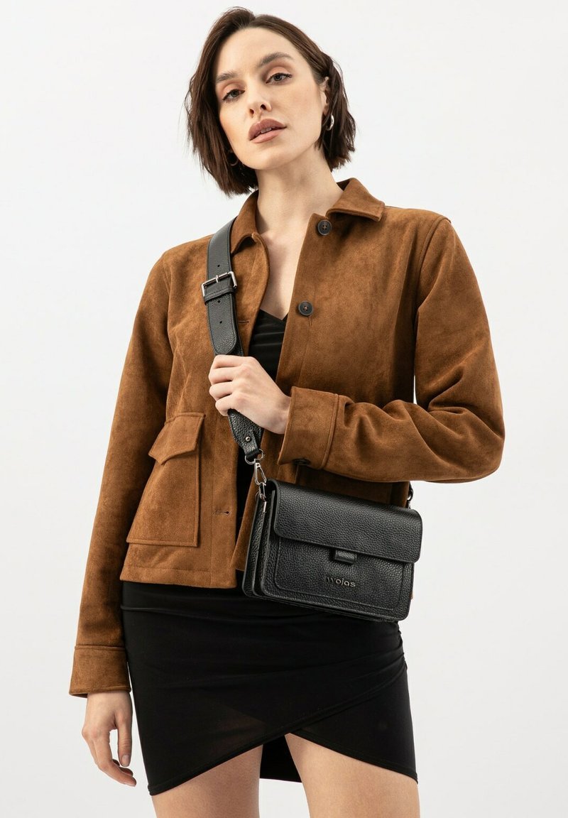 Schwarze Leder-Crossbody-Tasche mit strukturiertem Finish, magnetischem Klappenverschluss und verstellbarem Tragen, kombiniert mit einer braunen Suedjacke und einem schwarzen Kleid.