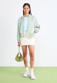 Mintgrön bomberjacka över ljusblå skjorta, beige kjol. Grön handväska med kedja, vita strumpor och ljuslila sneakers. Enhetlig bakgrund.