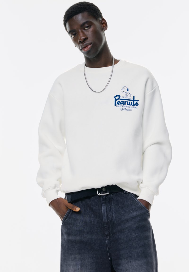 Witte sweatshirt met blauwe "Peanuts" graphic, geribbelde manchetten en een relaxed fit, gedragen met donkere spijkerbroek en een zilveren ketting.