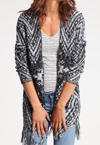 Cardigan en tricot noir et blanc avec un motif en chevrons, à ouverture frontale, à manches longues, et avec des détails en frange au niveau de l'ourlet. Porté au-dessus d'un haut rayé.