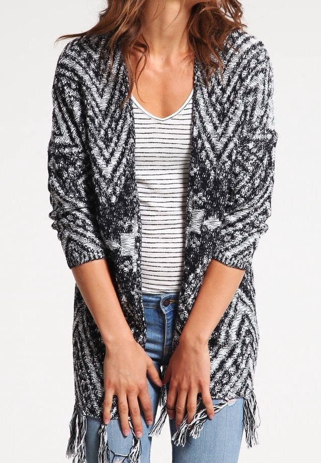 Cardigan en tricot noir et blanc avec un motif en chevrons, à ouverture frontale, à manches longues, et avec des détails en frange au niveau de l'ourlet. Porté au-dessus d'un haut rayé.