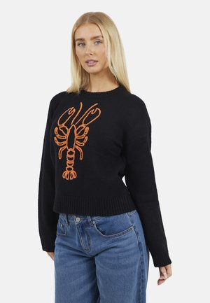 Donna con lunghi capelli biondi che indossa un maglione nero con un disegno arancione di un'aragosta e jeans blu, in piedi contro uno sfondo bianco.