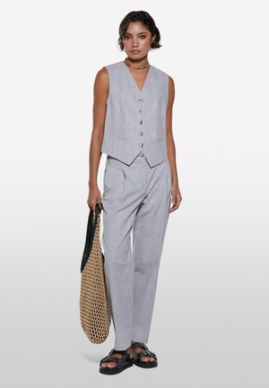 Gilet grigio e pantaloni coordinati realizzati in tessuto leggero, con scollatura a v, bottoni e pieghe, abbinati a una borsa intrecciata e sandali.