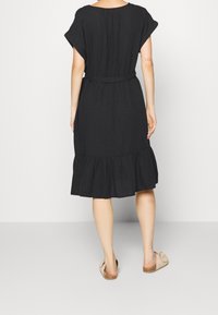 Vestido de lino negro con un corte holgado, mangas cortas, cintura ceñida con cinturón y bajo con volantes. Combinado con zapatos de lona beige.