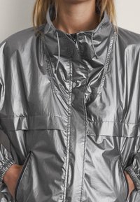 Frau in einer glänzenden silbernen Kapuzenjacke mit Reißverschluss, die von Hals bis Hüfte zu sehen ist, mit Händen in den seitlichen Reißverschlusstaschen.