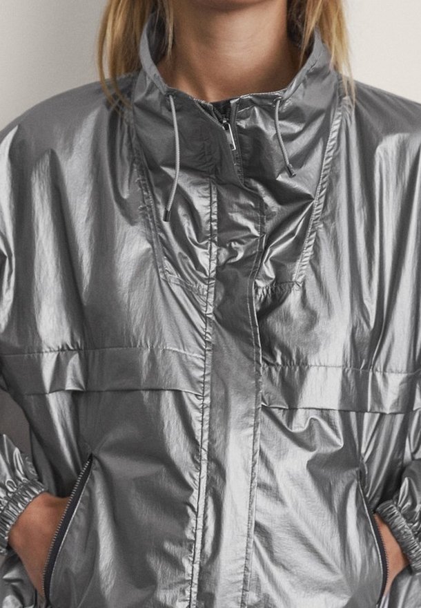 Donna che indossa una giacca a zip con cappuccio argentata lucida, con le mani nelle tasche laterali con zip, mostrata dalla testa ai fianchi.