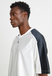 Camiseta deportiva blanca con acentos negros en las mangas, con un diseño de cuello en V y textura de malla, con un logo en la parte delantera.