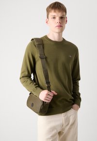 Olivgrüner Strickpullover mit Rundhalsausschnitt; leichtes Material. Helle Hose; rechteckige olivgrüne Schultertasche mit Logo.