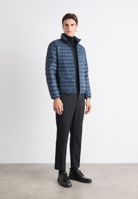 Giacca puffer blu navy con zip frontale e colletto alto, caratterizzata da un design trapuntato orizzontale, indossata sopra un dolcevita nero e abbinata a pantaloni neri.