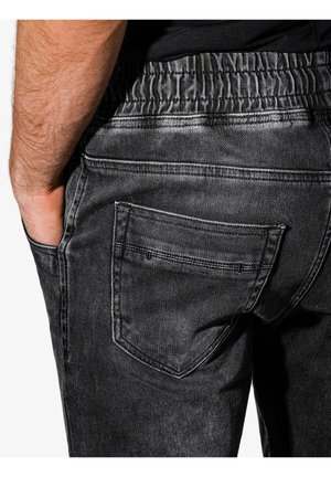 Mann trägt schwarze Denim-Jeans mit elastischem Bund und Hand in der linken Vordertasche, gezeigt von der Taille bis zur Mitte des Oberschenkels.