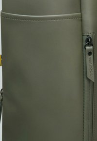 Olive green backpack with smooth leather texture, featuring a side zip pocket and branded tag. Minimalist design with clean lines.