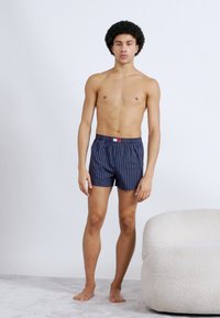 Μπόξερ σε ναυτικό μπλε με κάθετες ρίγες, ελαστική μέση, μικρή κόκκινη και λευκή ετικέτα, κατασκευασμένα από ελαφρύ ύφασμα, με ομαλή υφή.