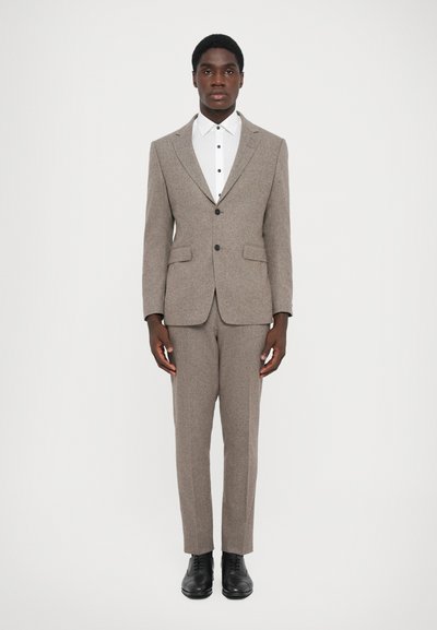 Costume en tweed gris avec une coupe ajustée, veste à un bouton, et revers crantés. Chemise blanche avec des boutons noirs en dessous. Chaussures noires.