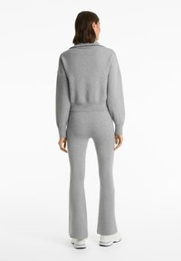 Vrouw gezien van achteren in een lichtgrijze, lange mouw, crop sweater en bijpassende flared broek met witte sneakers op een witte achtergrond.