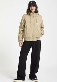 Beige hooded jacket with drawstrings, front zipper, and side pockets, paired with black wide-leg pants and black sneakers.