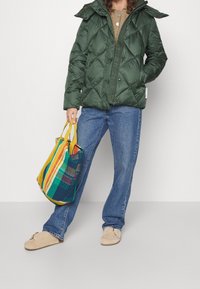 Grøn quilted pufferjakke med hætte, parret med blå jeans og beige slippers. Holder en flerfarvet stribet vævet totebag.