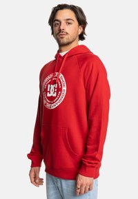 Red hoodie made of cotton blend, featuring a front pocket, ribbed cuffs, and large printed logo on the chest. Smooth texture.