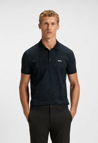 Svart polotröja med ett texturerat, geometriskt mönster; har krage och korta ärmar; liten vit logotyp på bröstet.