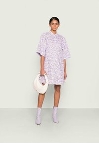 Vestido de manga curta em roxo claro com padrão floral amarelo, apresentando um colarinho abotoado e um corte solto. Combinado com uma mala branca e botas lilás.