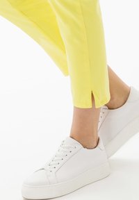 Zapatillas blancas de cuero con suelas planas y diseño con cordones, combinadas con pantalones amarillos recortados que tienen aberturas laterales en el dobladillo.